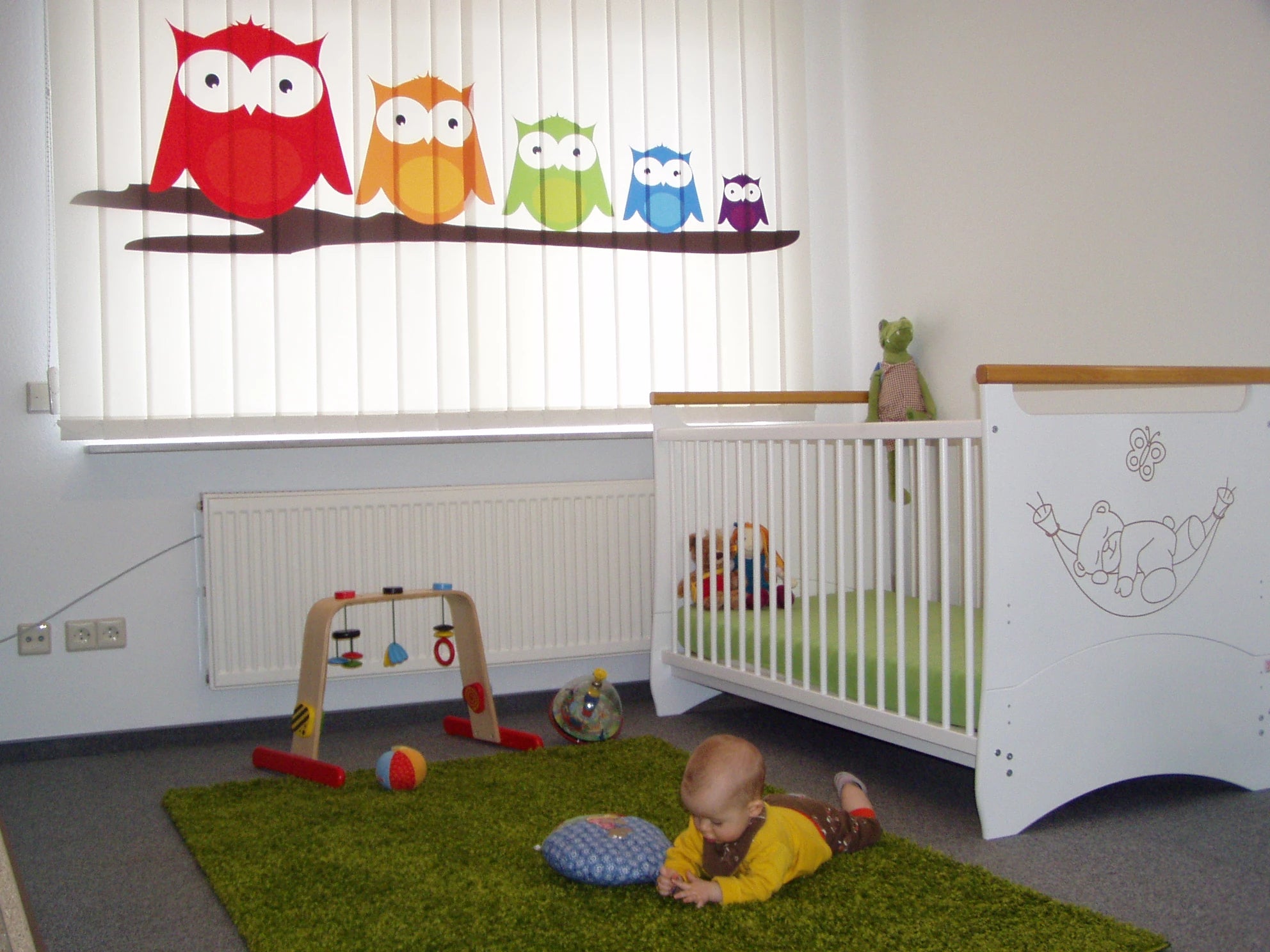 bunte Eulen gedruckt auf einem Foto-Lamellenvorhang für ein Kinderzimmer, in dem ein Baby friedlich spielt