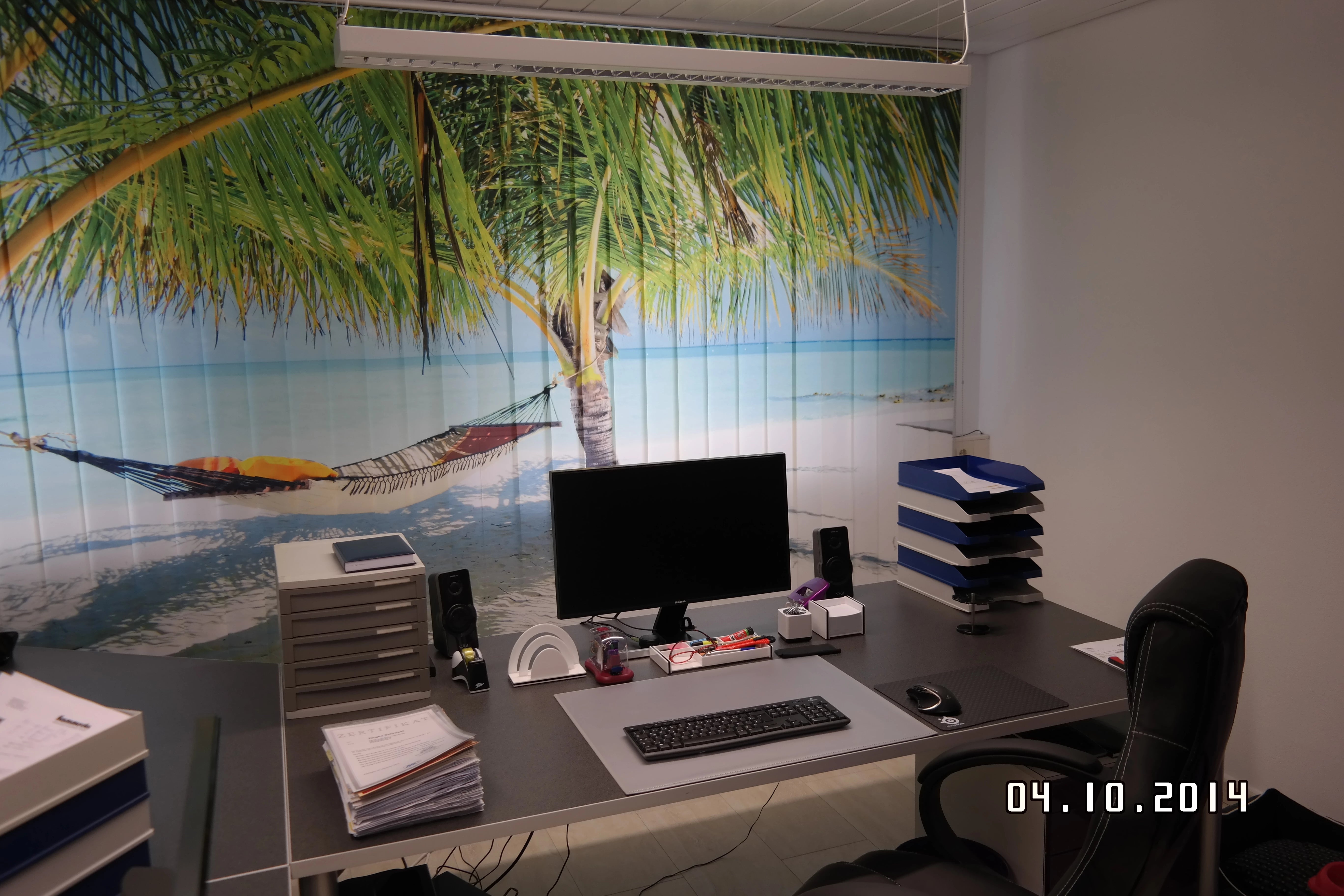 ein Büro mit Fensterfront, vor dem ein großer mit Palmen bedruckter Lamellenvorhang am Strand hängt für ein bisschen Urlaubsfeeling und zum träumen