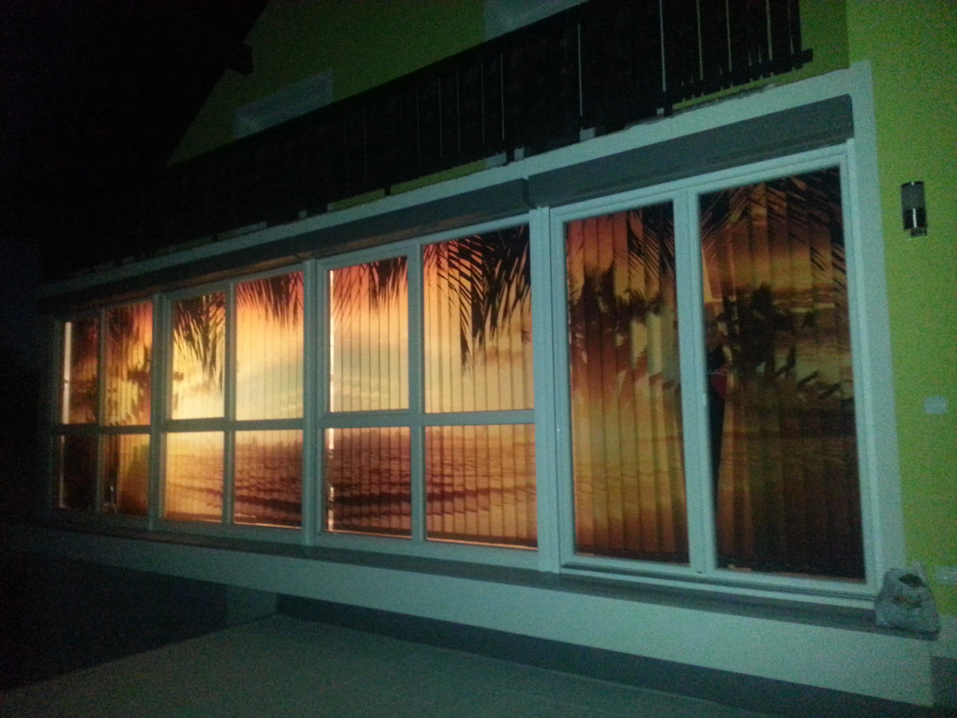 Ansicht beleuchtete Rückseite von einem großen bedruckten Foto-Lamellenvorhang an einer großen Fensterfront bedruckt mit Palmen bei Sonnenuntergang am Meer