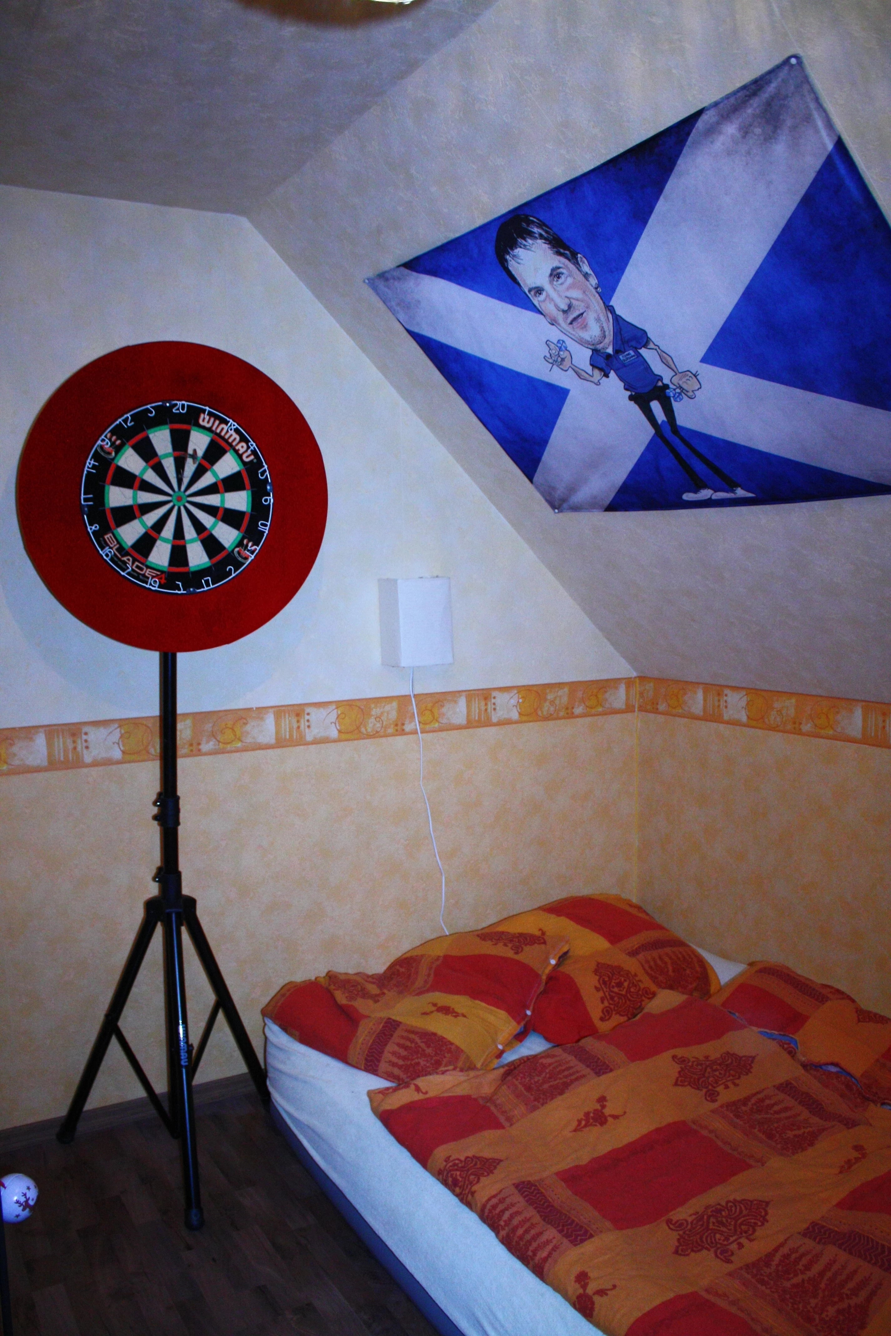 mit Karikatur des Darts-Spieler Gary Anderson bedruckte Foto-Fahne an der Dachschräge neben der Dart-Scheibe im Schlafzimmer 