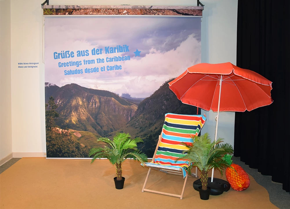 Fotorollo als Fotohintergrund bedruckt mit einer Berglandschaft mit Grüßen aus der Karibik einer Ausstellung im botanischen Garten in Berlin mit Liegestuhl, Palme und Sonnenschirm für Besucher, um sich fotografieren zu lassen 