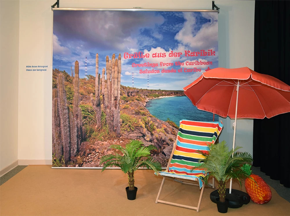 Fotorollo als Fotohintergrund bedruckt mit einem Bild von Kakteen und der Küste und Grüßen aus der Karibik einer Ausstellung im botanischen Garten in Berlin mit Liegestuhl, Palme und Sonnenschirm für Besucher, um sich fotografieren zu lassen 
