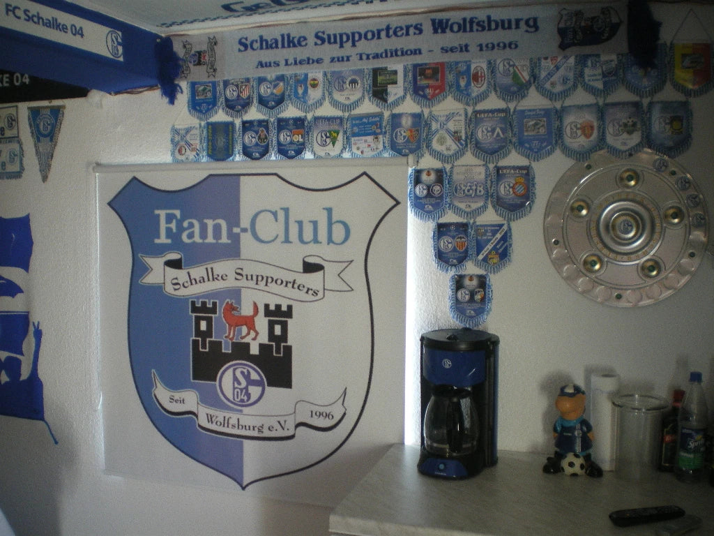 ein Club-Raum der Fan-Supporter in Wolfsburg für den 1. FC Schalke 04 mit einem mit Logo bedruckten Foto-Rollo am Fenster eines mit Fan-Artikel geschmückten Raum