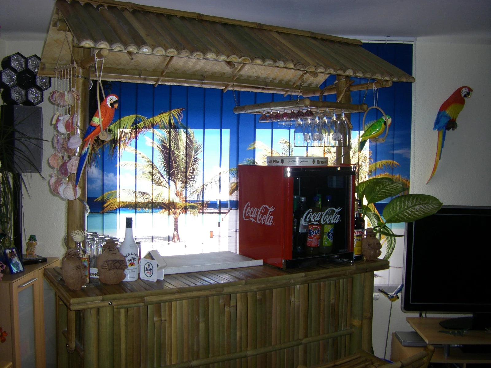 Eine im Wohnzimmer aufgebaute Bar, hinter der ein Lamellenvorhang mit einem Strandmotiv aus der Südsee mit Palmen hängt