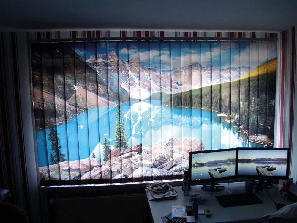 Ein Büroschreibtisch mit zwei Monitoren. Dahinter ein Fenster, das von einem Lamellenvorhang bedeckt ist. Der Lamellenvorhang zeigt ein Motiv mit einem klaren, blauen Bergsee