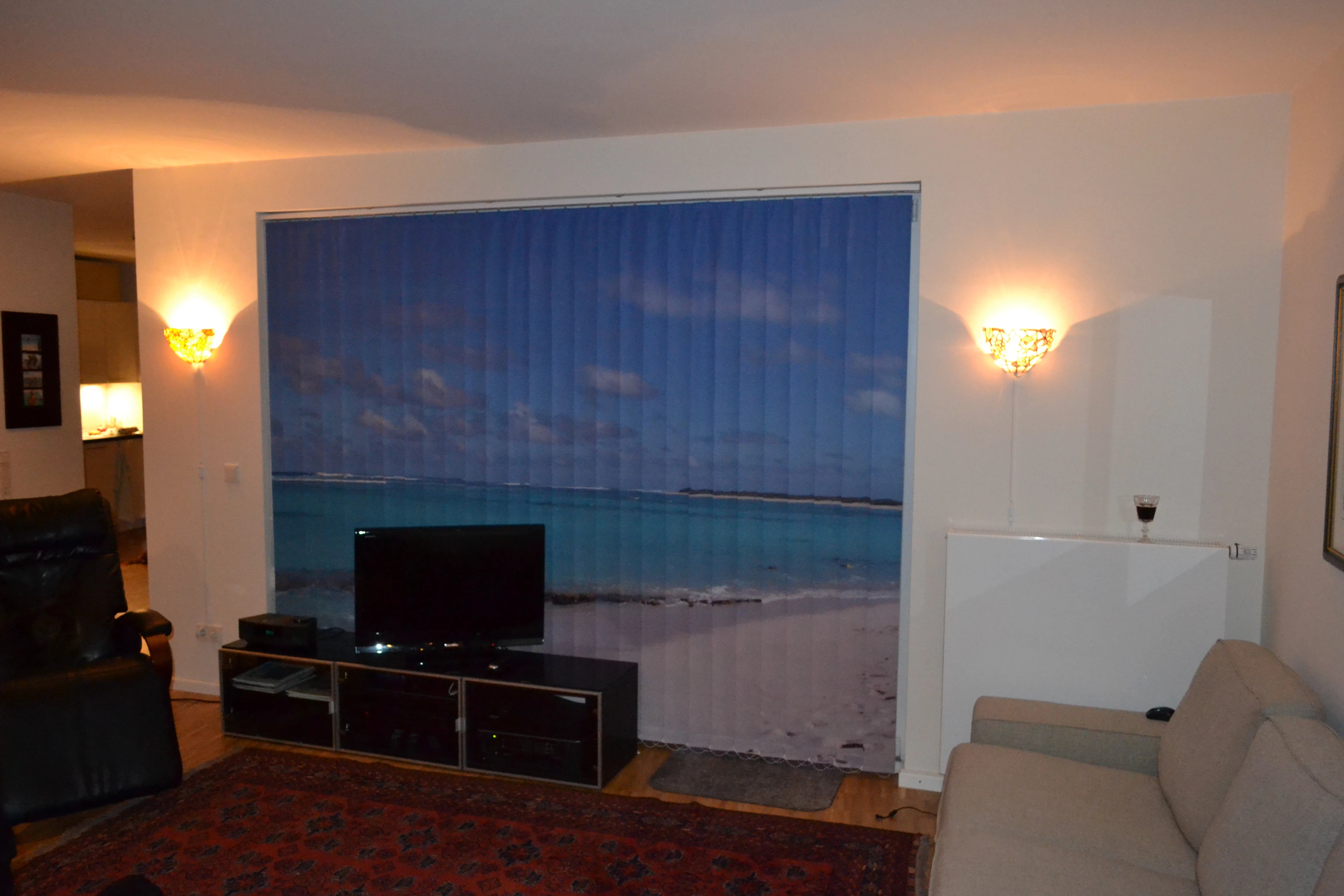 Ein klassisch eingerichtetes Wohnzimmer mit Sofa und Sessel. Auf einem Sideboard steht ein Fernseher. Das dahinter liegende Fenster wird mit einem Lamellenvorhang abgedeckt, das ein Strandmotiv aus der Südsee zeigt