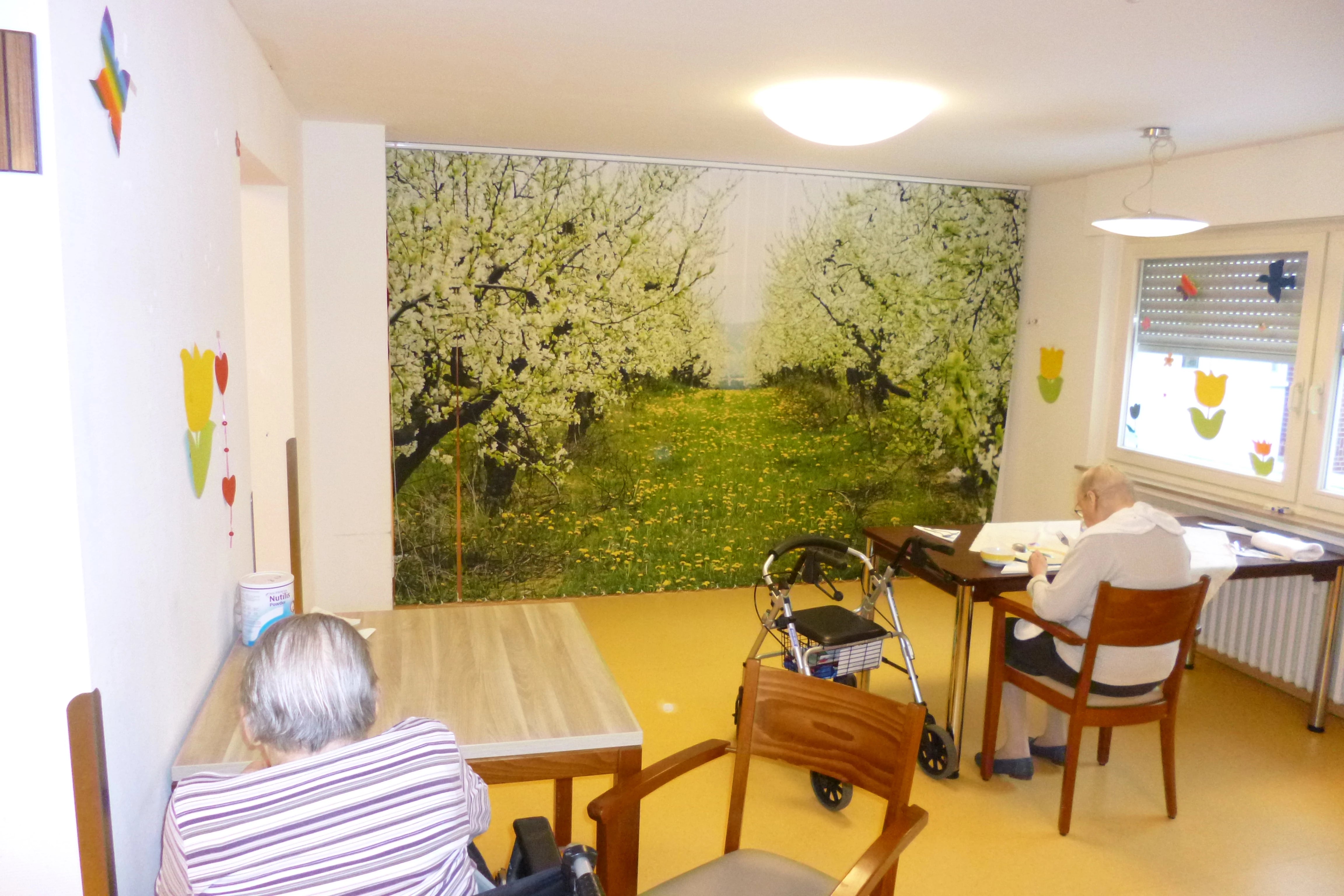 Im Aufenthaltsraum eines Pflegeheimes hängt ein mit Apfelbäumen bedruckter Lamellen-Vorhang