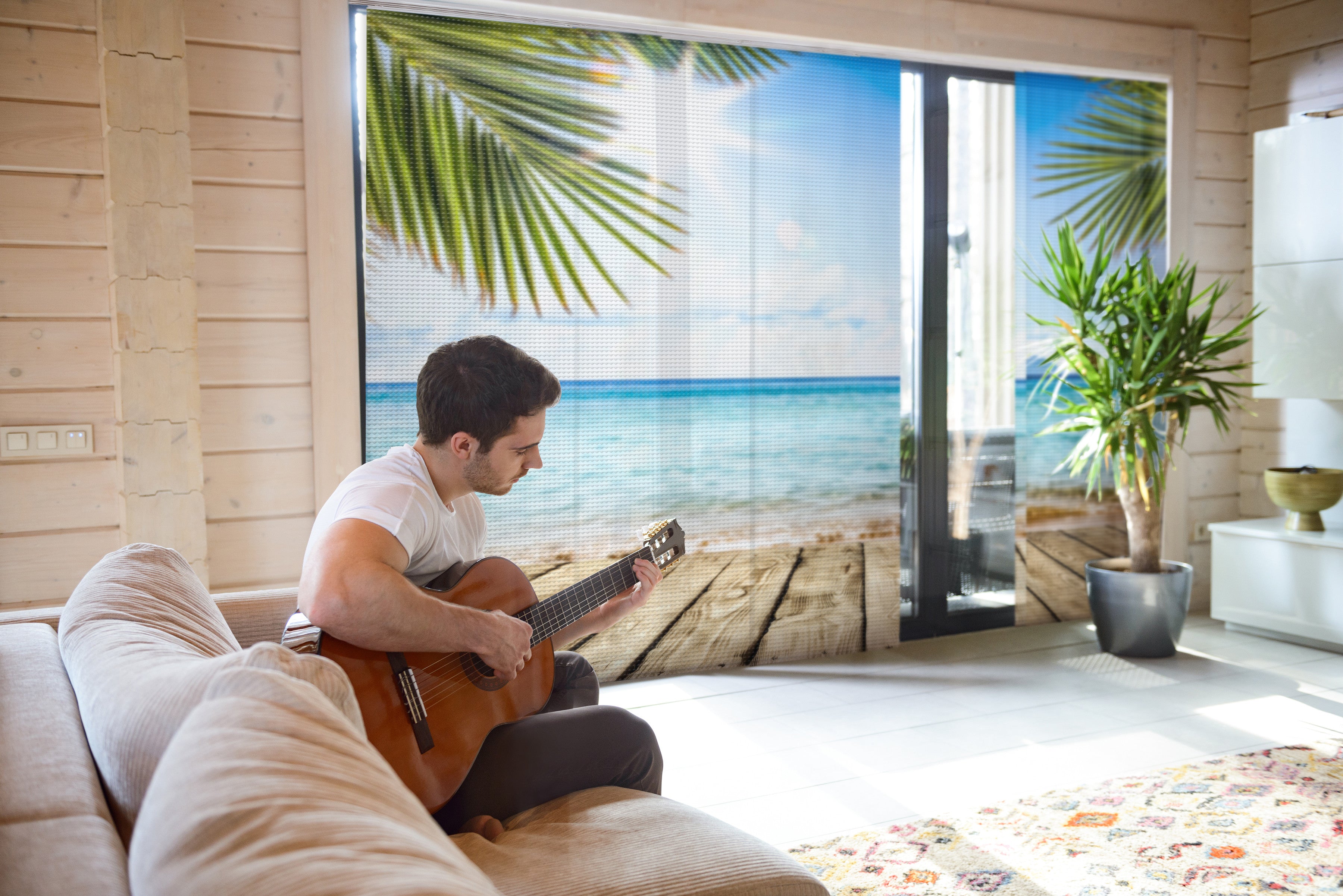 Ein junger Mann sitzt auf der Couch und spielt Gitarre. Im Hintergrund eine große Fensterfront mit Akustik-Schiebegardinen, bedruckt mit einen Meer und Palmenmotiv